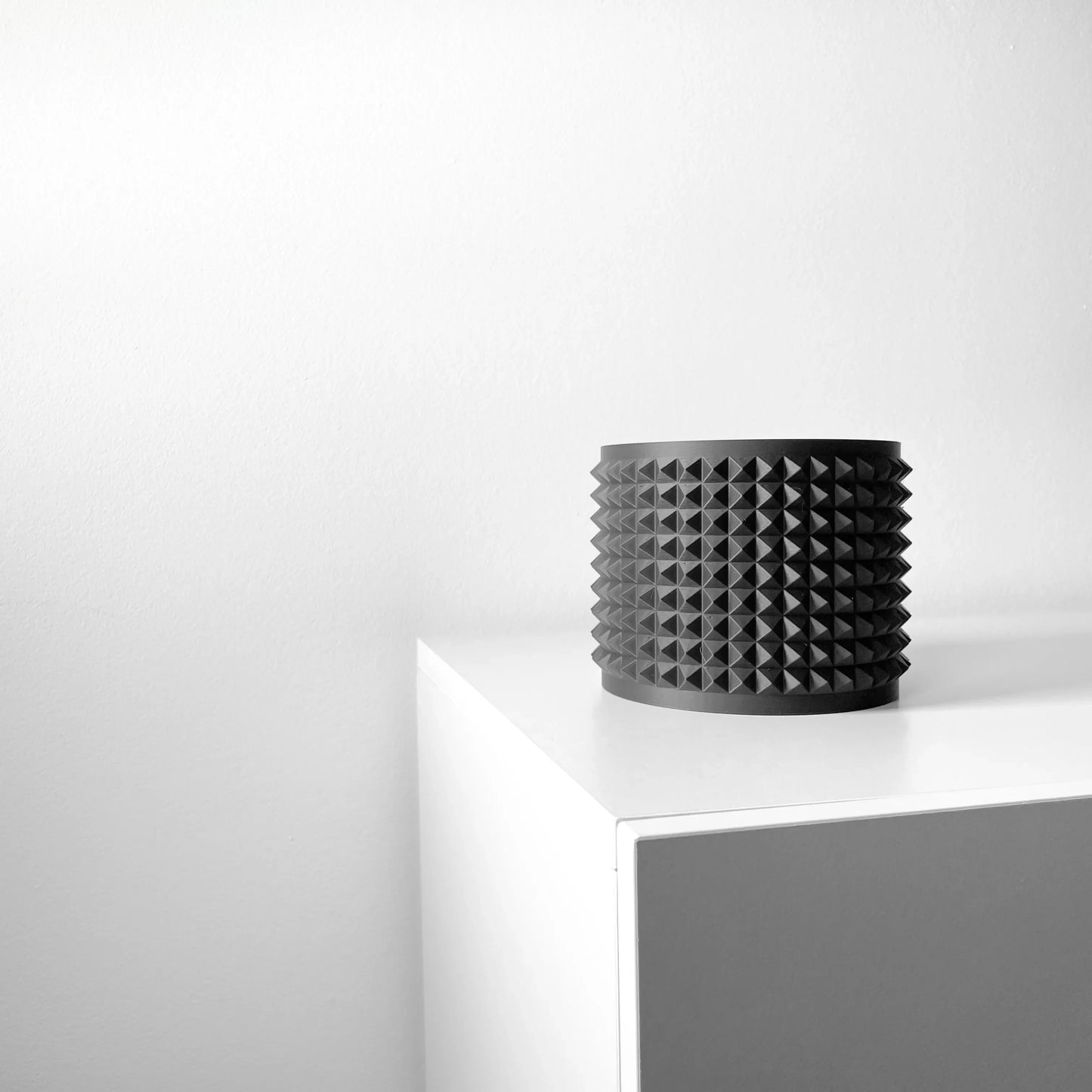 Pot décoratif cylindrique noir mat avec surface texturée en picots uniformes sur un meuble blanc minimaliste.