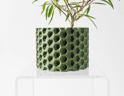 Pot décoratif vert olive cylindrique avec motif hexagonal en relief, posé sur un support transparent.