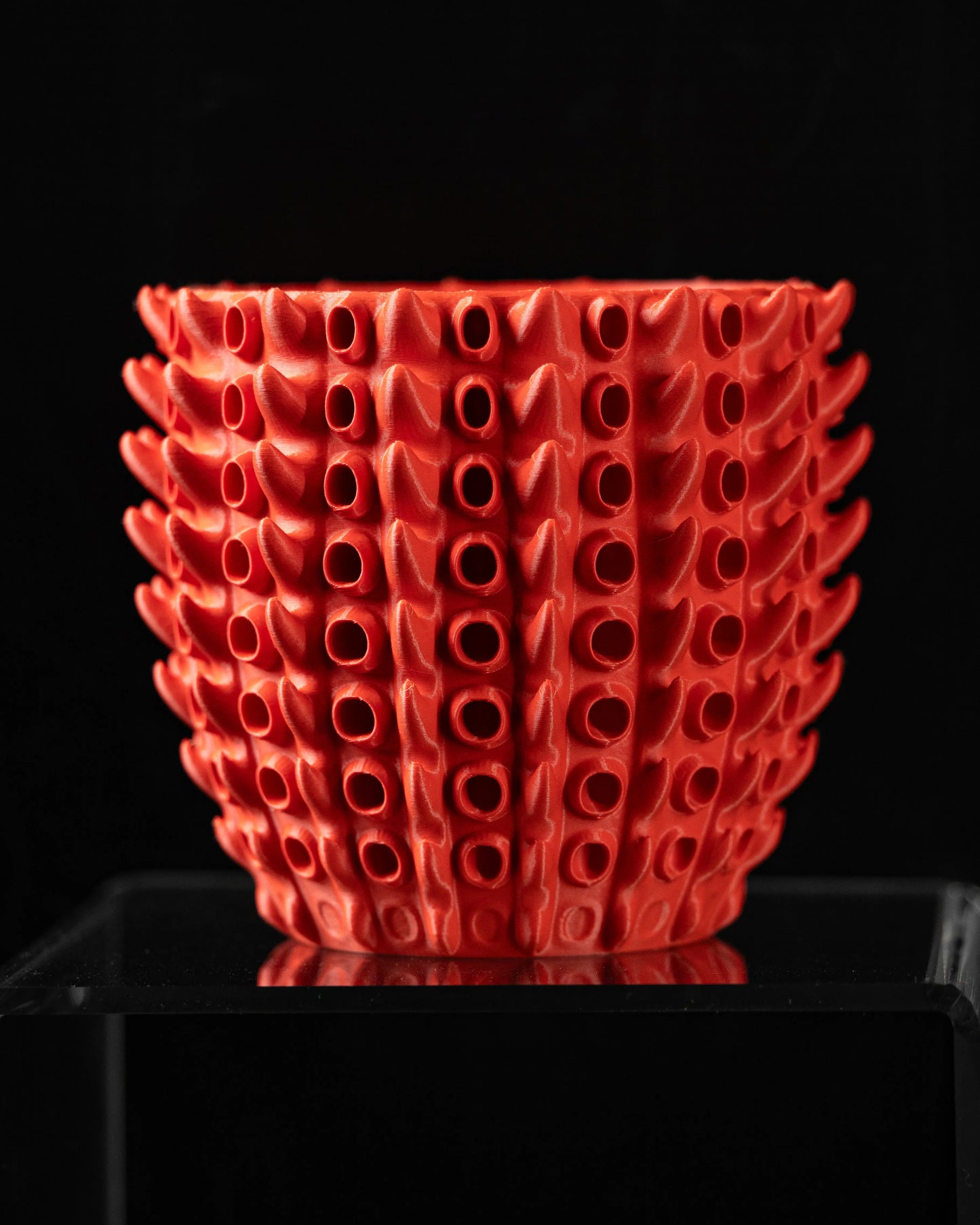 Pot décoratif rouge vif avec surface texturée composée de rangées de petites ouvertures ovales en relief, forme arrondie.