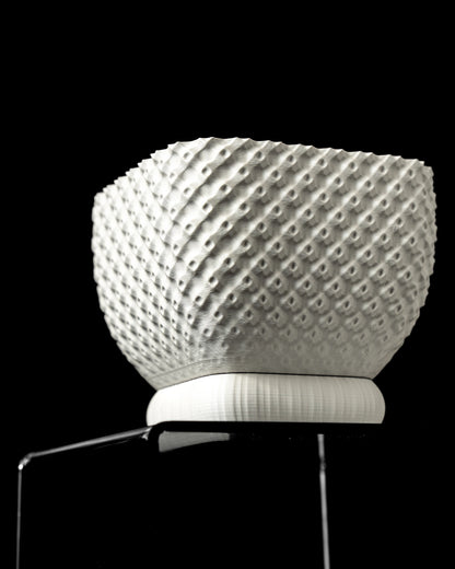 Pot décoratif blanc en matière texturée à motifs géométriques en relief, forme arrondie et base plate sur un support.