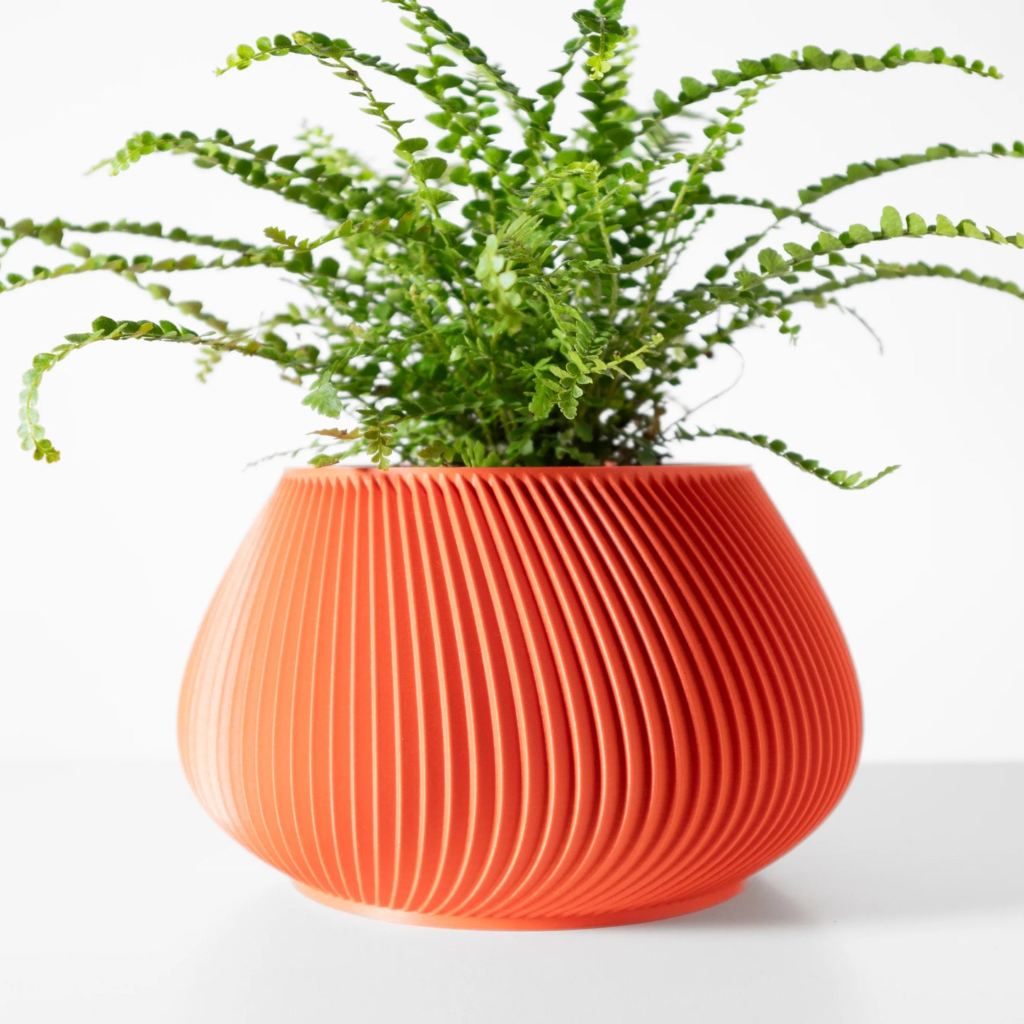 Pot décoratif orange vif en plastique moderne avec rainures verticales incurvées et fougère verte luxuriante à l’intérieur.