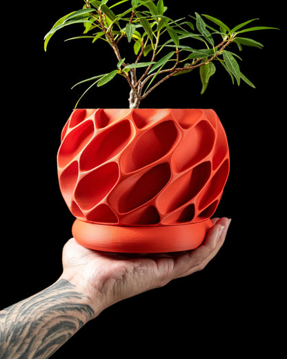 Pot décoratif rouge en plastique moderne avec surface texturée en forme de nid d'abeille, posé sur une main tatouée, plante.