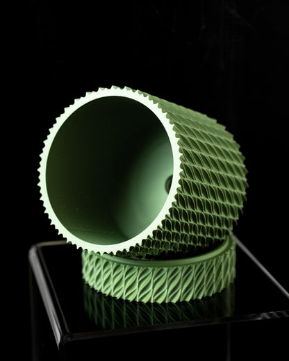 Pot décoratif vert clair cylindrique incliné, surface extérieure texturée en relief géométrique, intérieur lisse, posé sur.