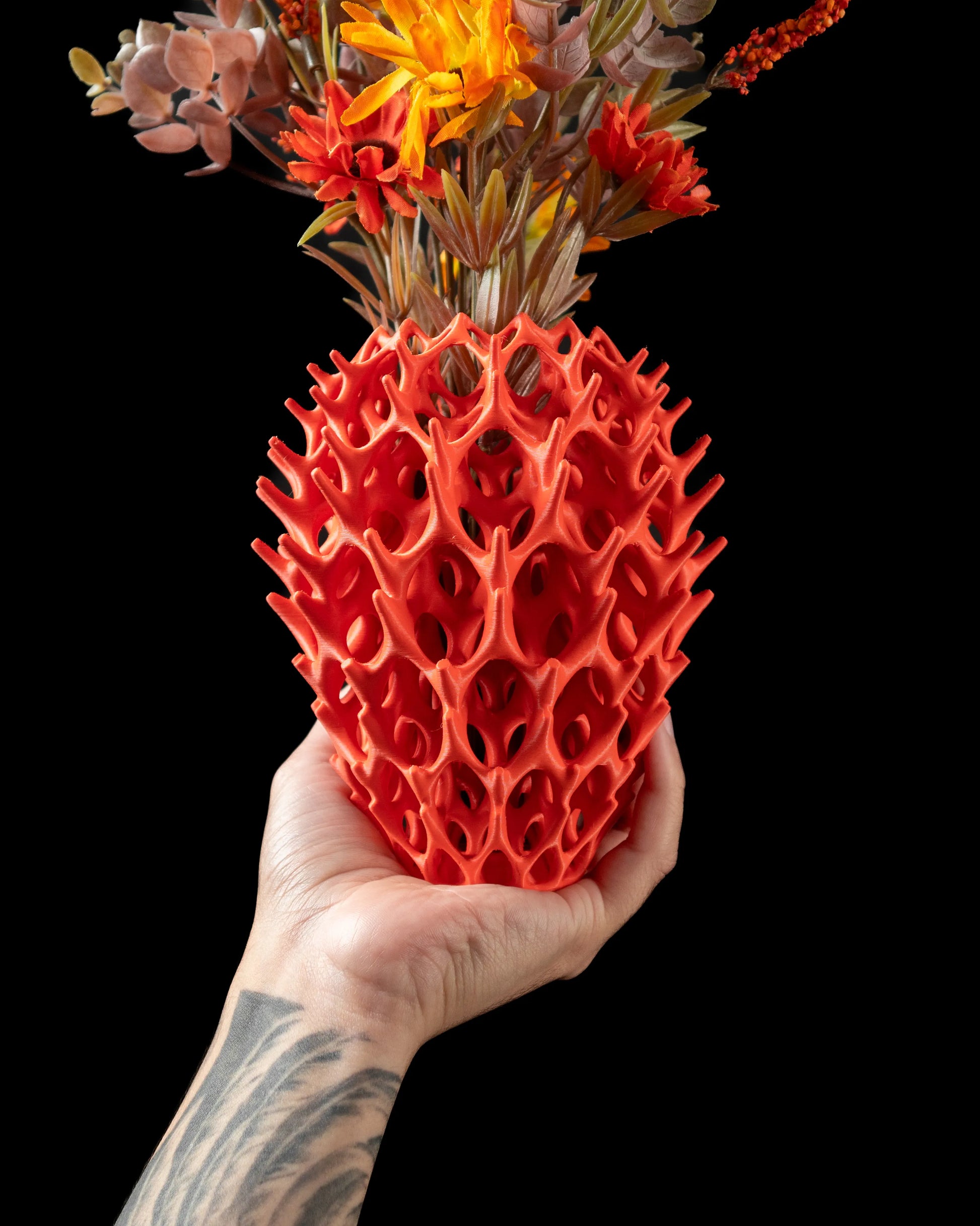 Vase rouge vif avec surface texturée en forme de bulles allongées, tenu par une main tatouée, contenant des fleurs séchées.