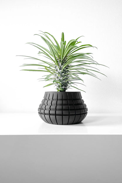Pot décoratif noir mat en forme de sphère aplatie avec motifs carrés en relief, planté d’une plante verte à longues feuilles