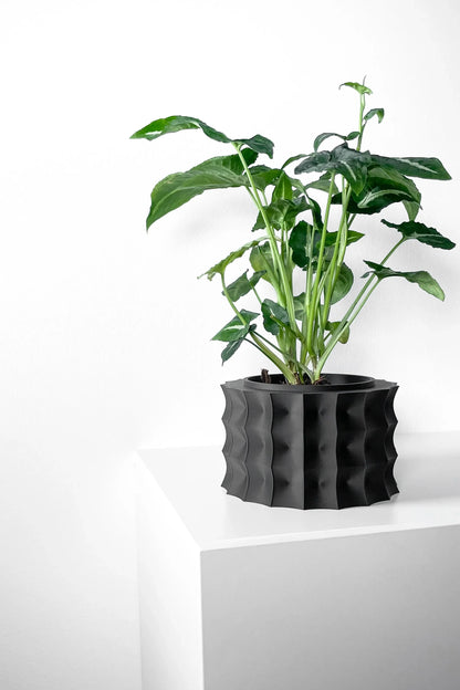 Pot décoratif noir mat avec texture ondulée et plante verte aux larges feuilles posée sur surface blanche.