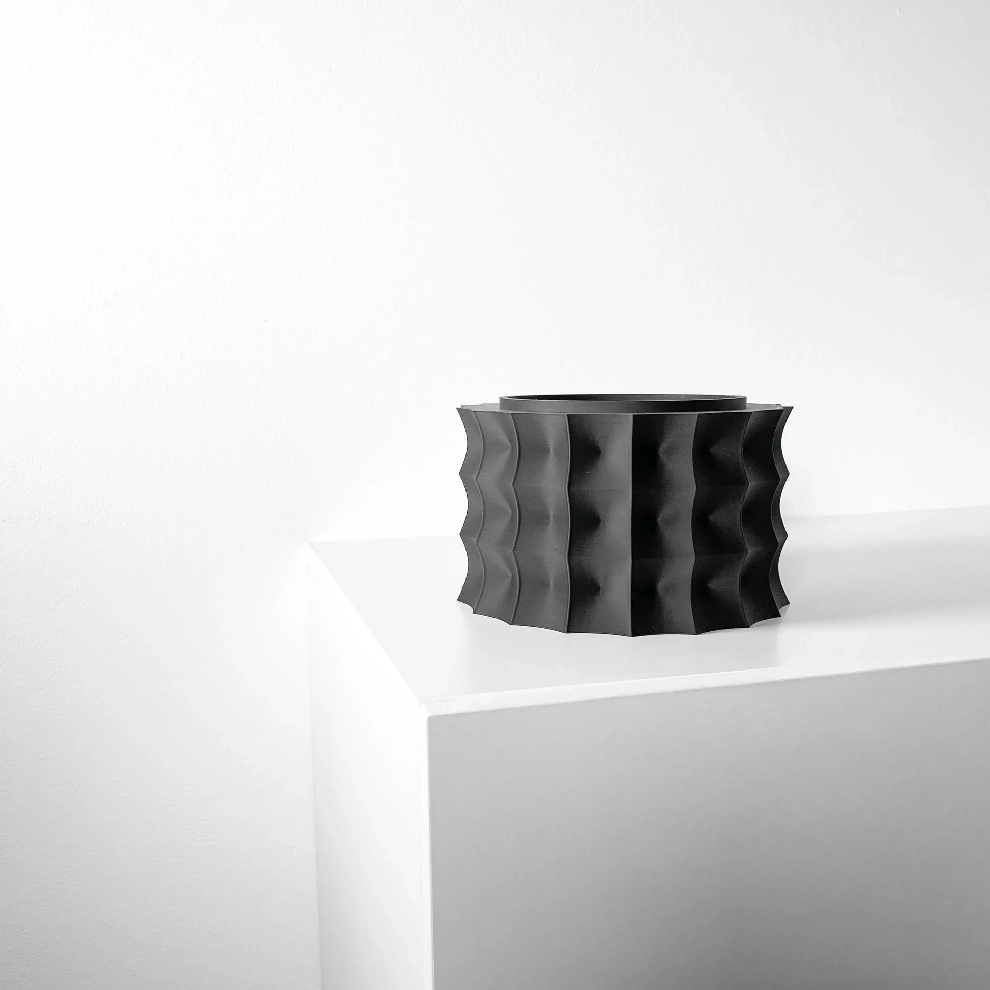 Pot décoratif noir mat avec surface ondulée en relief, forme cylindrique large et moderne, posé sur un coin blanc.