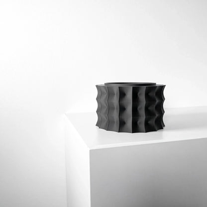 Pot décoratif noir mat avec surface ondulée en relief, forme cylindrique large et moderne, posé sur un coin blanc.
