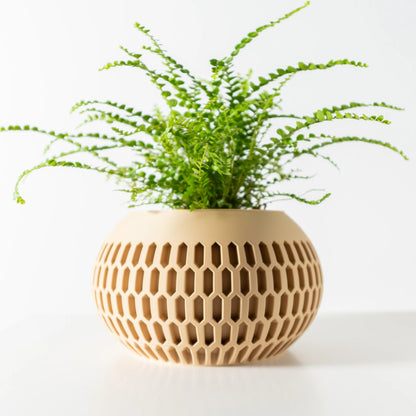 Pot décoratif rond en matériau beige avec motif hexagonal ajouré, plante verte suspendue à l’intérieur.