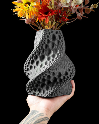 Vase noir mat en forme de spirale texturée avec des empreintes circulaires, tenu à la main contenant des fleurs séchées.