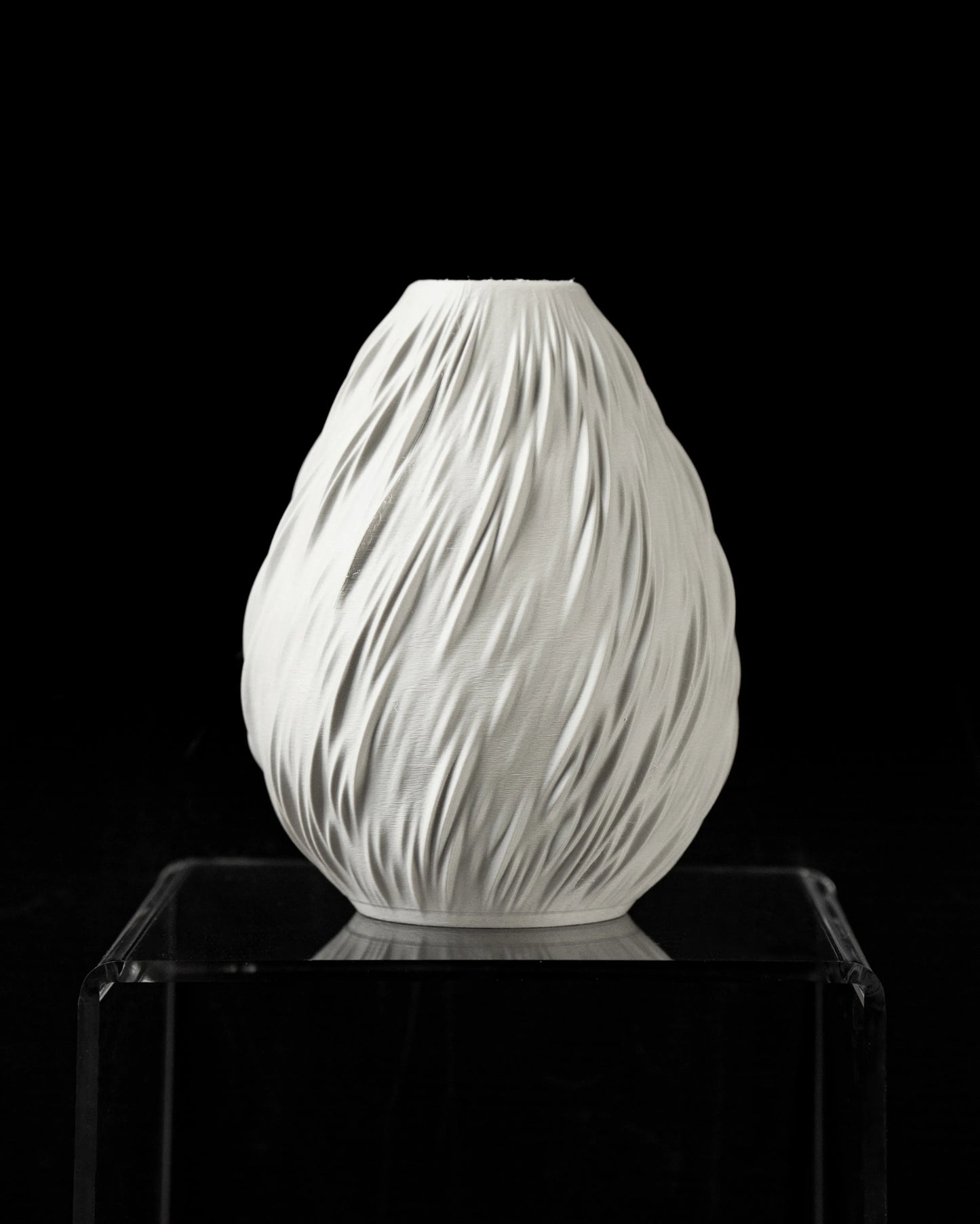 Vase blanc mat en forme ovale avec texture ondulée verticale rappelant des gouttes de pluie, posé sur un support transparent.