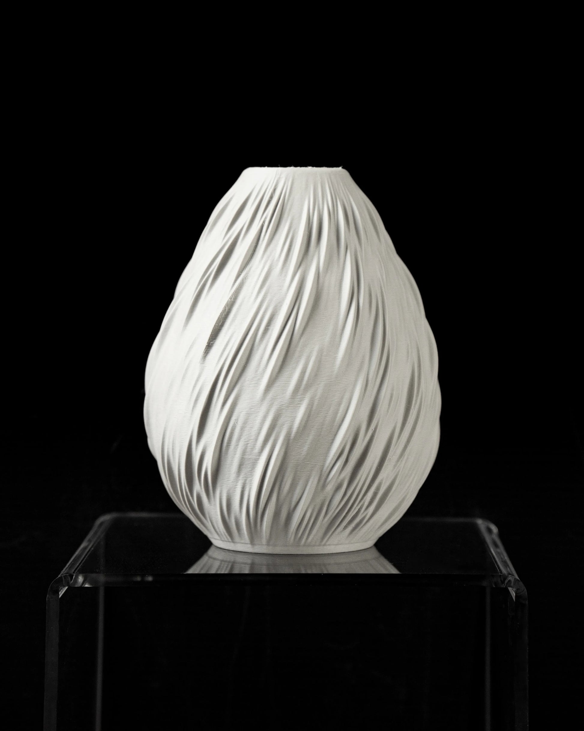 Vase blanc mat en forme ovale avec texture ondulée verticale rappelant des gouttes de pluie, posé sur un support transparent.